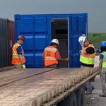 Workers in safety vests and helmets inspecting a blue shipping container on a flatbed trailer.