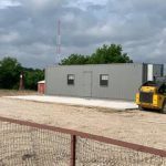 A small gray building and a yellow skid-steer loader on a gravel lot with a fence in the foreground and trees in the background.