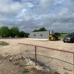 Construction site with a portable building, vehicles, and machinery, enclosed by a metal fence on a cloudy day.