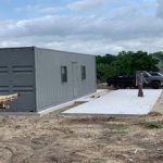A gray container office on a concrete foundation with a person standing nearby and a truck in the background at a construction site.