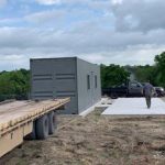 A person stands near a prefabricated structure on a concrete foundation beside a flatbed trailer, with trees in the background.
