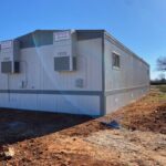 A white and gray trailer on dirt ground with trees in the background.